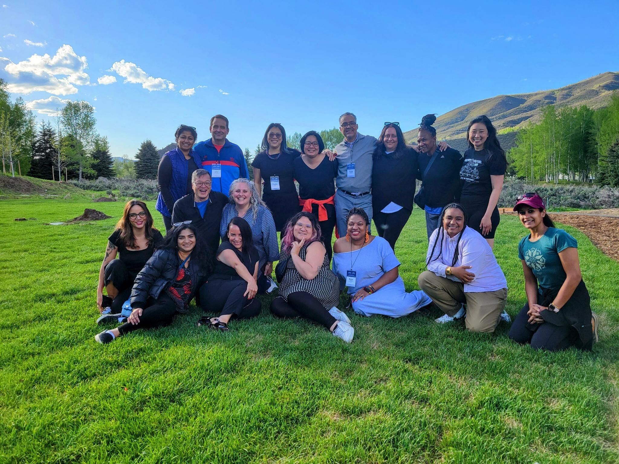 Group of individuals sitting and standing in front of a grassfield with mountains and blue skies in the back