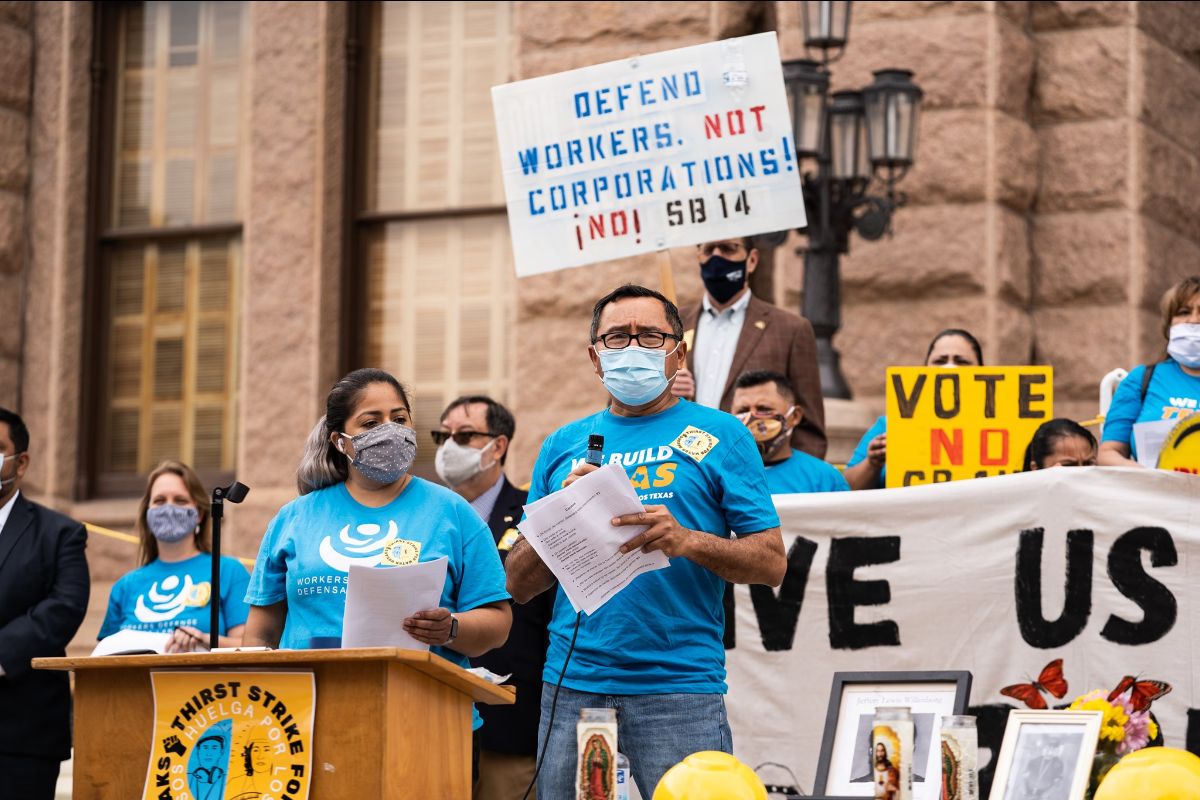 A photo of a group of a protestors wearing shirts from Workers Defense Project, standing at a podium and carrying a sign that says, 