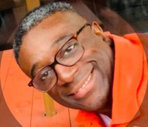 Photo of Gregory Wilson smiling, wearing glasses and an orange top