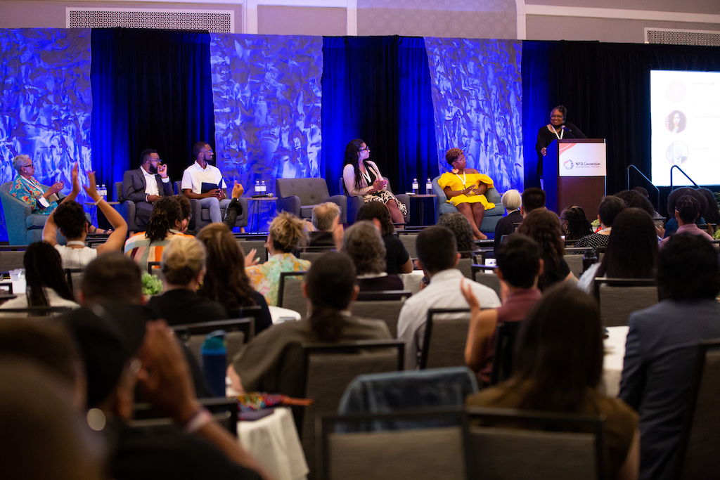 Photo of Chi, Chris, Curtis, Darlene, La'Meshia, and Sue on stage for the "Welcome to Wilmington! The Roots of Racial Capitalism in Eastern North Carolina and the South" plenary in front of a crowd full of seated attendees.