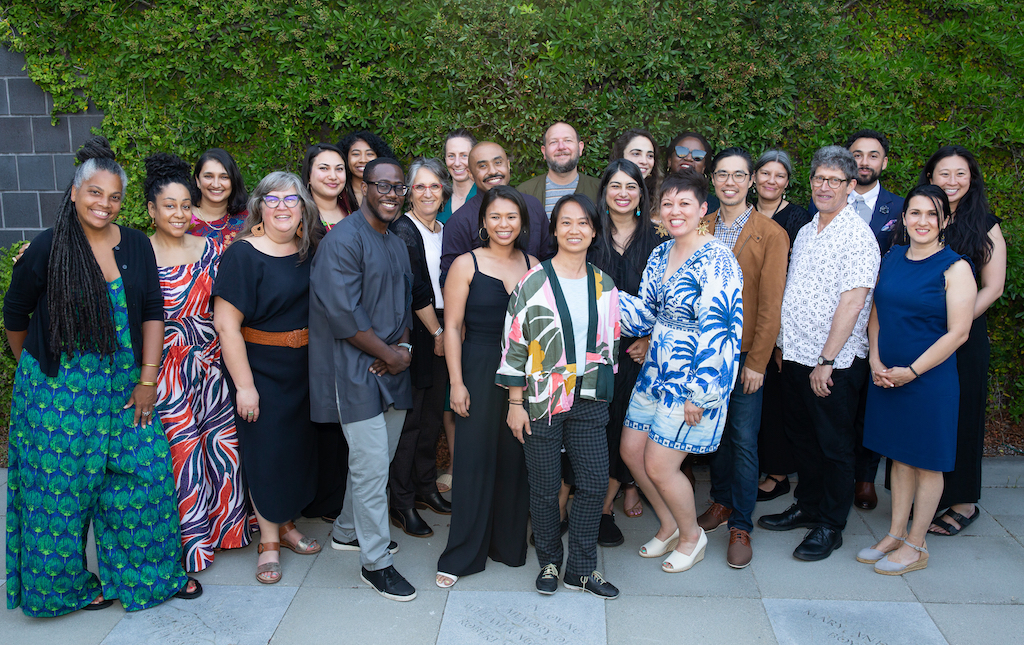 A group photo of NFG staff and board smiling in front of a brick wall covered in lush greenery.