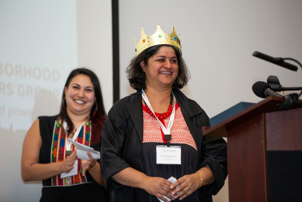 Shona stands smiling with a gold crown behind a podium. Neda Said stands behind her smiling and clapping.