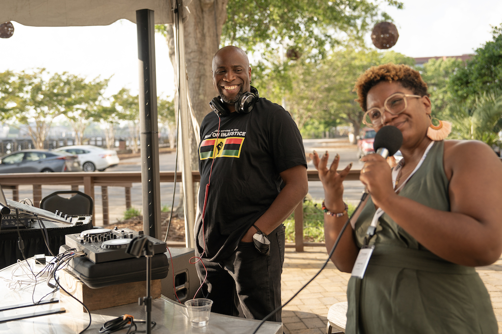 Nat and Chi stand behind a DJ set up. Nat has headphones around his neck and is smiling at Chi who is speaking into a microphone.