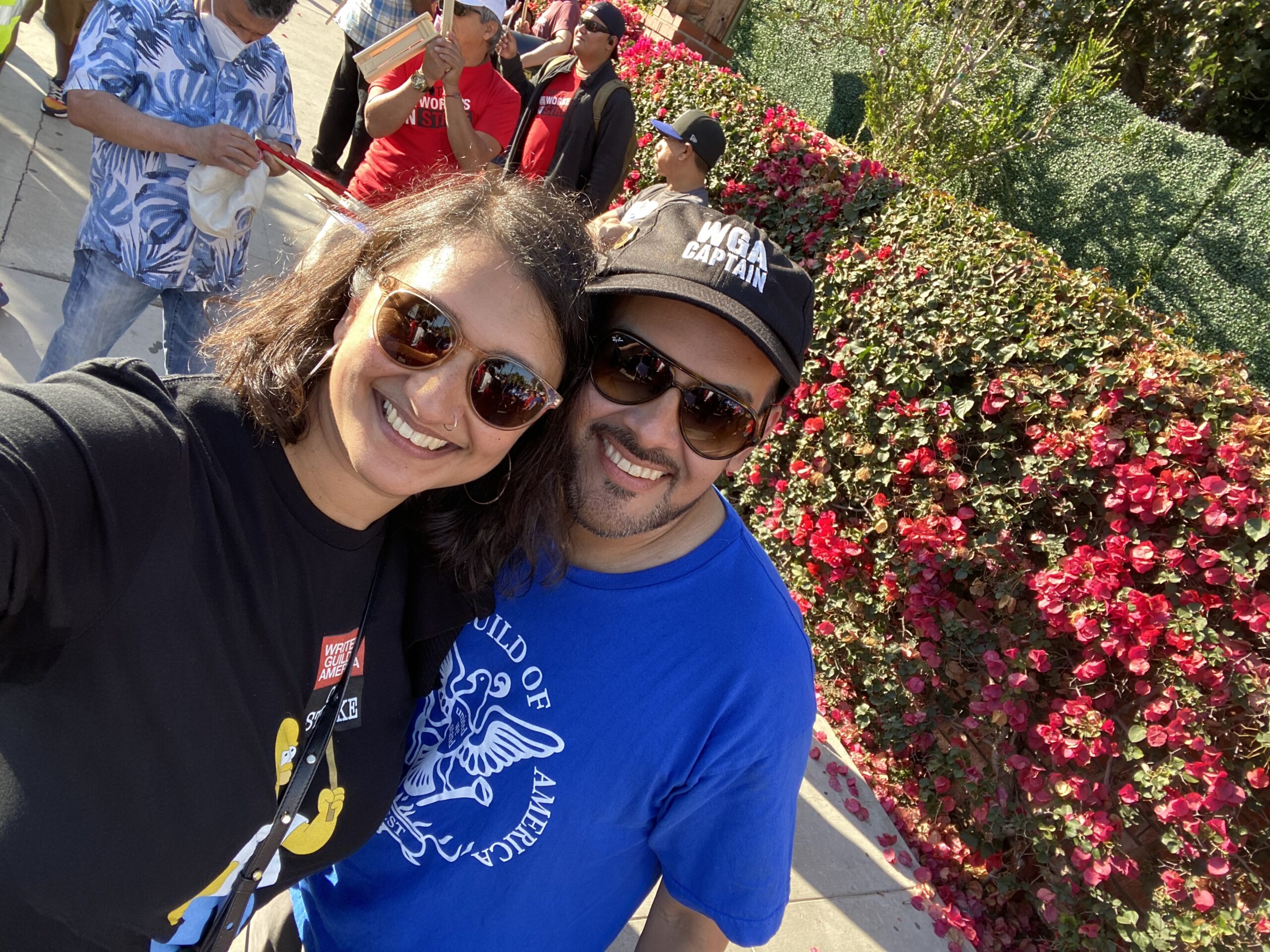 Manisha and her partner take a selfie in front of a flower bush on workers on strike in Los Angeles, CA. Manisha is wearing a black graphic t-shirt and sunglasses. Her partner is wearing a blue graphic t-shirt, sunglasses, and a black embroidered baseball cap that says WGA Captain.