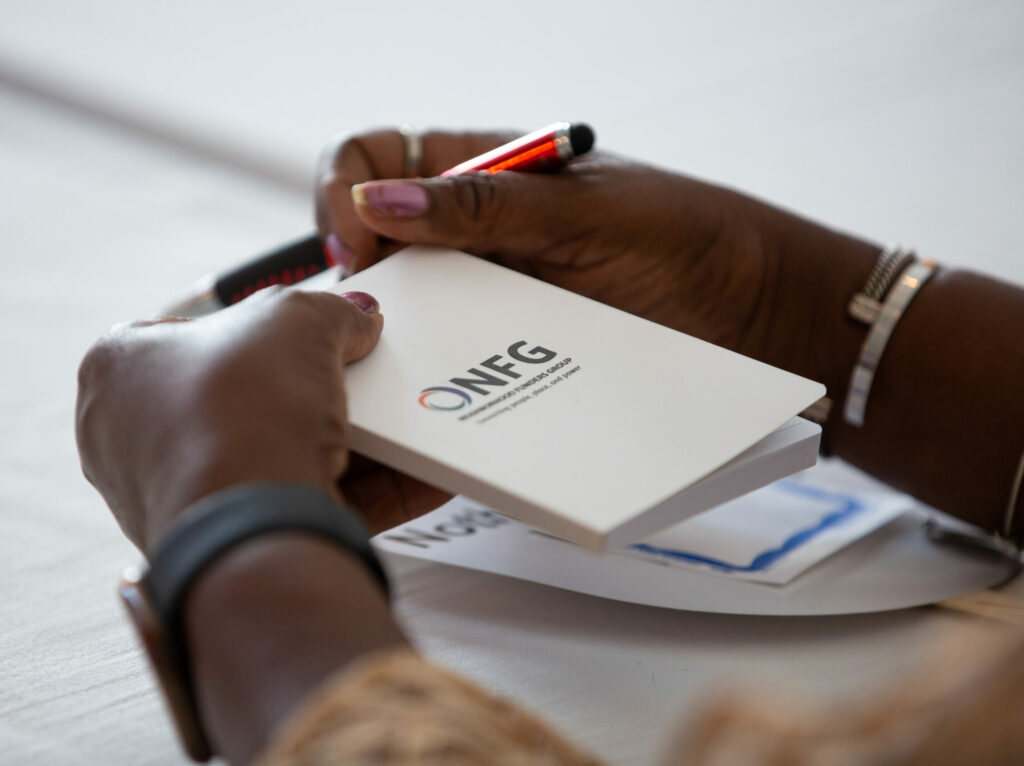 A close-up photo of two hands holding a small, white NFG branded notebook. The person holding the notebook also has a pen in their hands. They are wearing a watch on their right hand and two bracelets on the left hand.