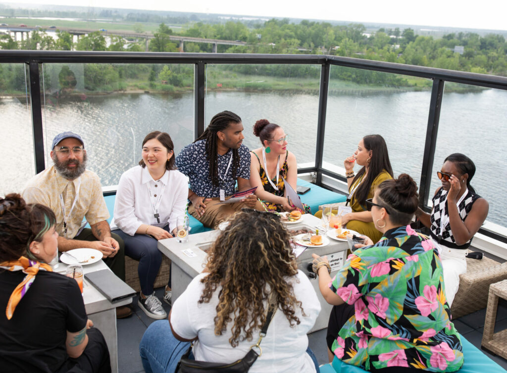 A group of funders gathered on at a rooftop restaurant during NFG's 2023 National Convening. Funders are conversing and sitting around a large table. In the background is a river.