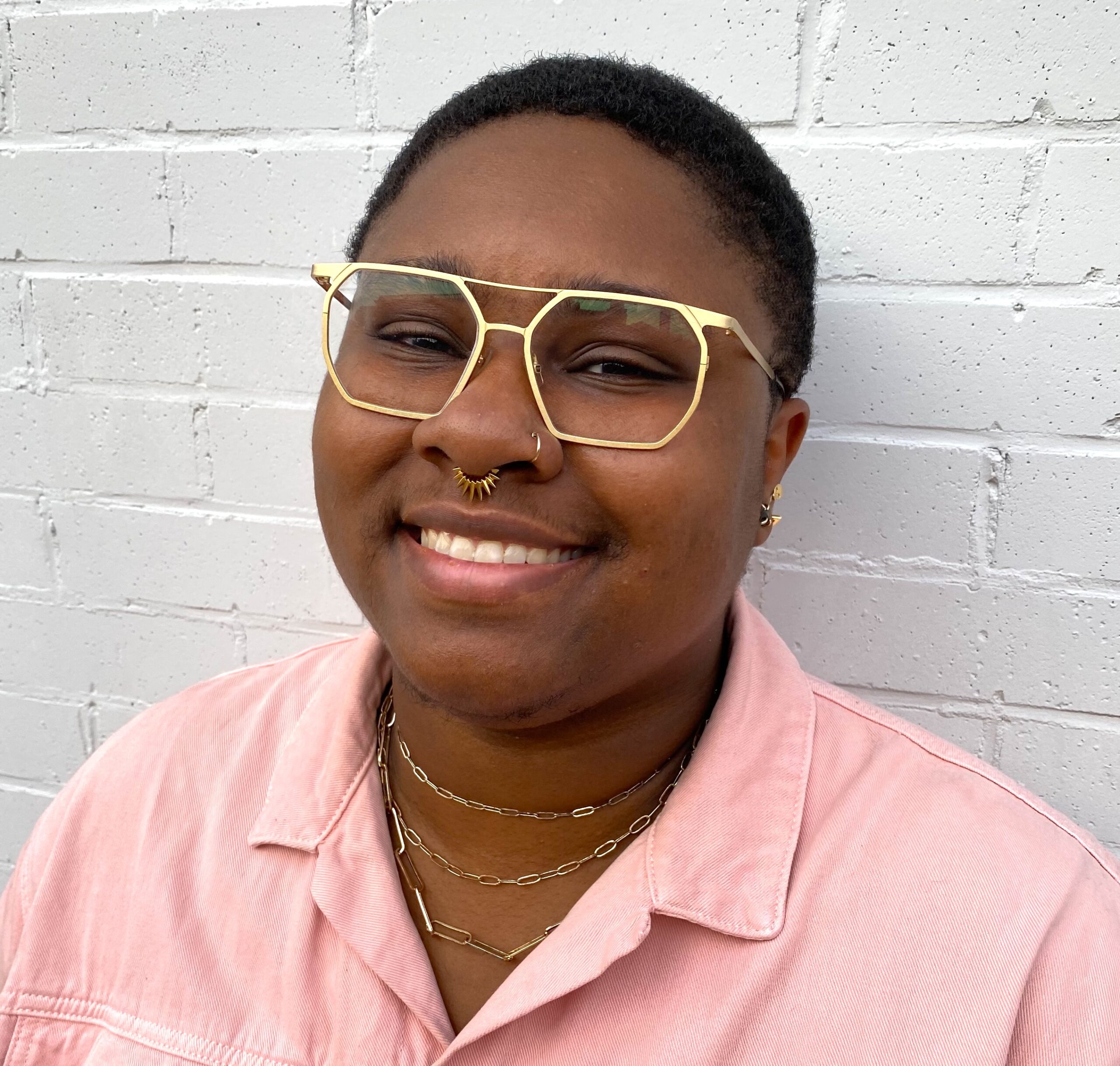 Headshot for Tahirah Green. Tahirah is smiling in front of a white brick wall, and wears a pink button-up shirt with gold rim glasses.