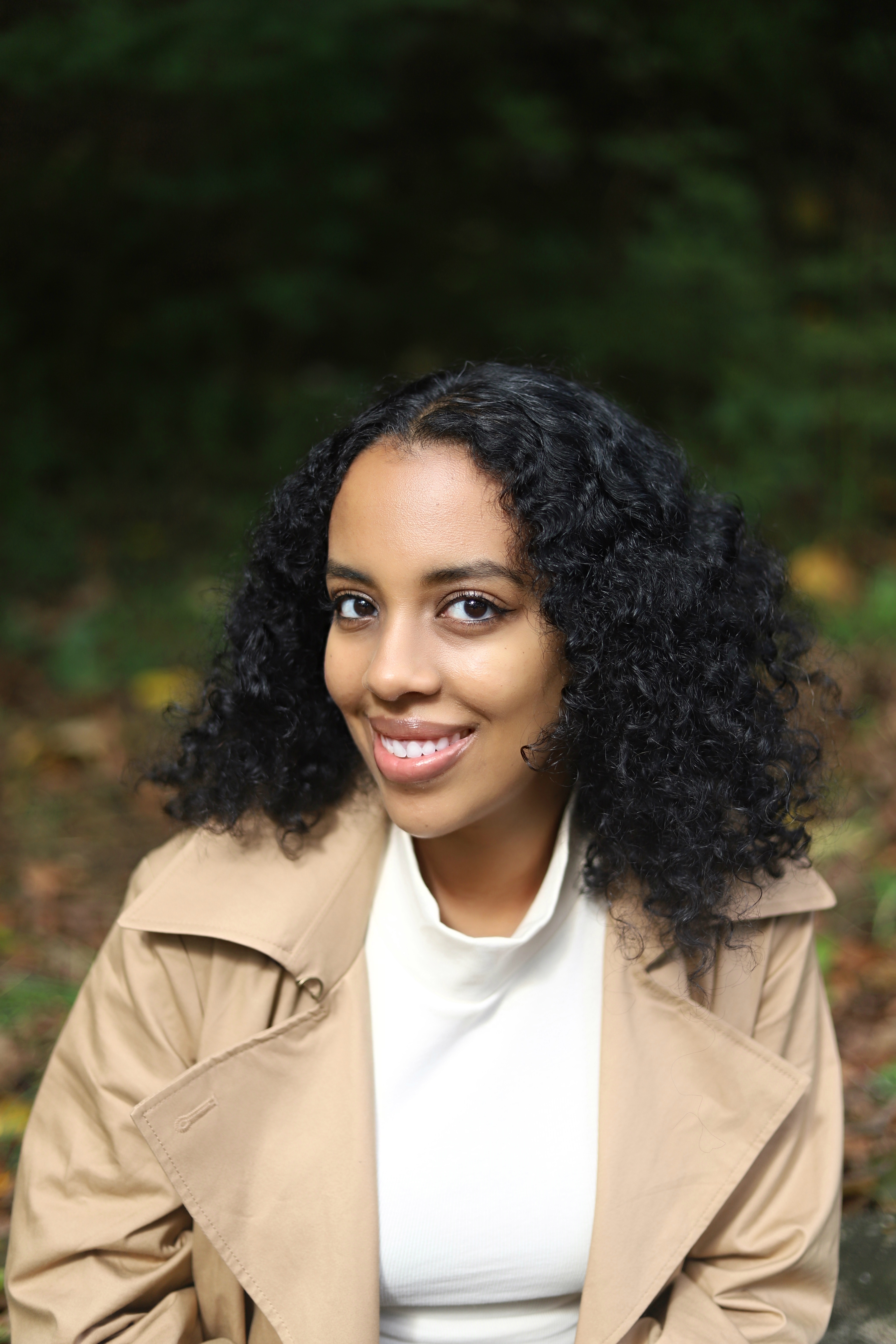 Headshot of Rahel Tekola, head turned to the side but gaze forward and smiling in a white shirt and tan jacket.