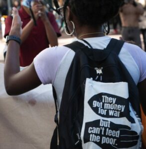 Photo from behind a person with a backpack on, with a pinned screenprinted quote on fabric that reads, 'They got money for war but can't feed the poor -Tupac'